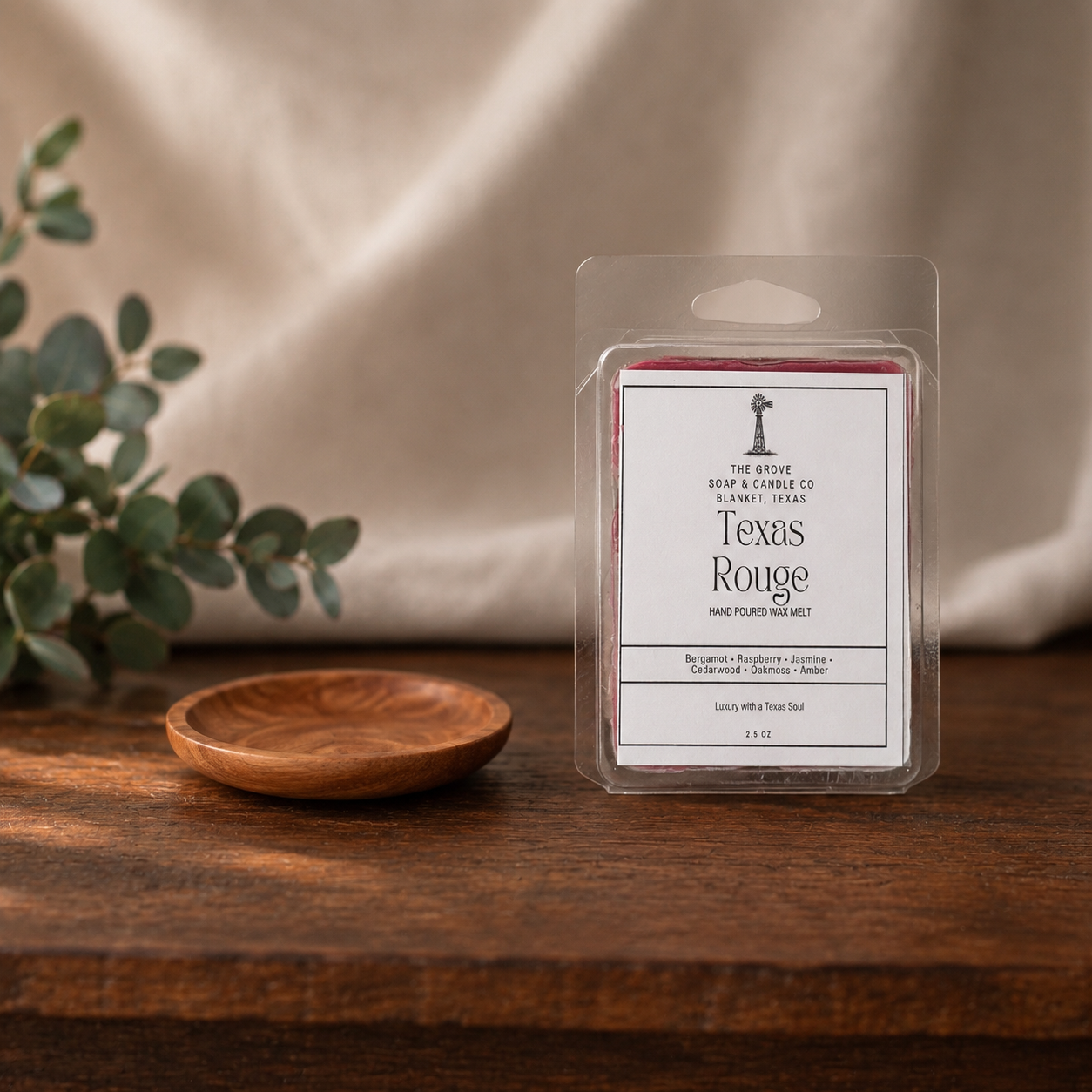A packaged wax melt product labeled Texas Rouge by The Grove Soap & Candle Co. stands on a wooden surface against a draped beige fabric backdrop, with eucalyptus leaves and a small wooden dish nearby.