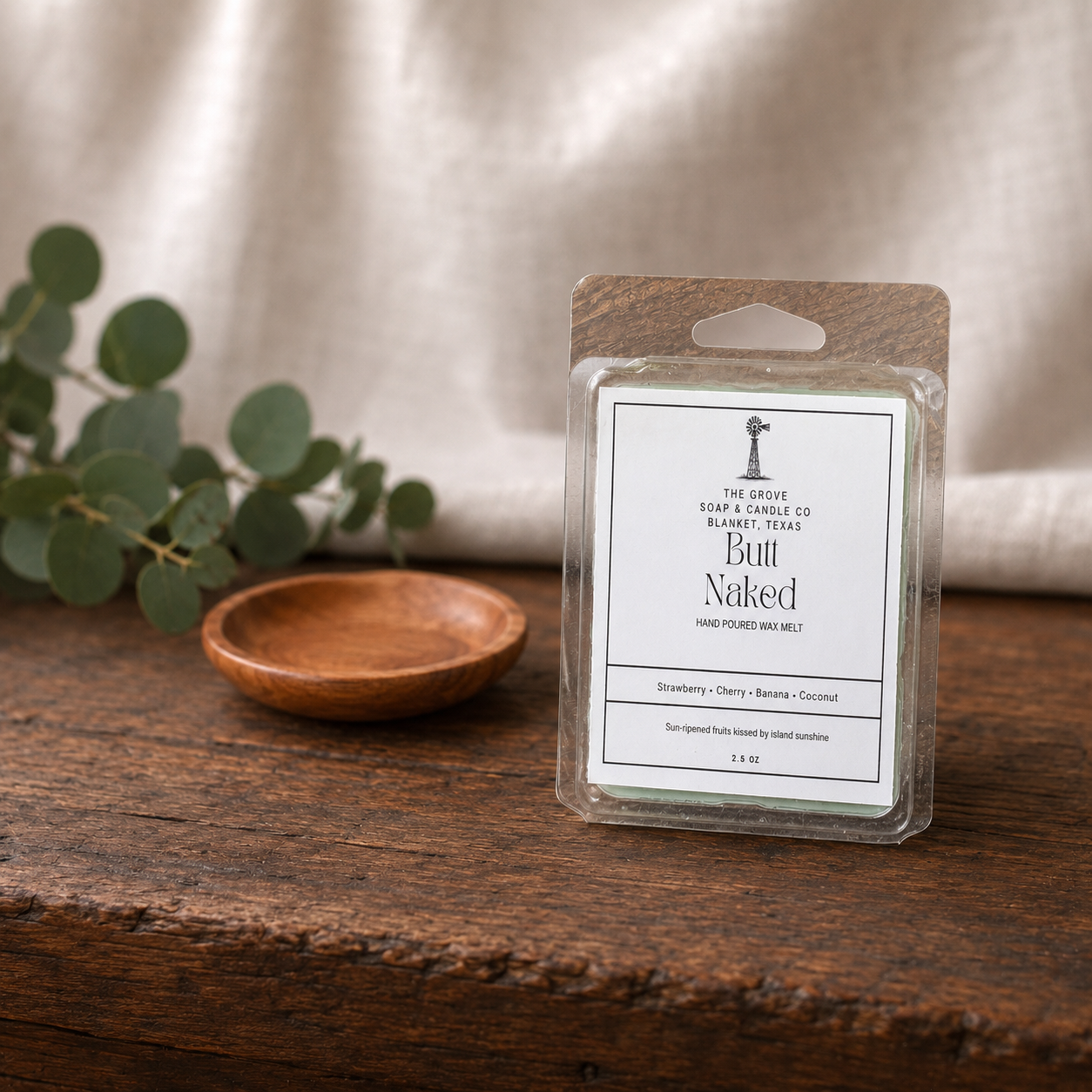 A packaged wax melt product labeled 'Bull Naked' by The Grove Soap and Candle Co sits on a rustic wooden surface with a small wooden dish and eucalyptus leaves in the background against a soft neutral fabric backdrop.