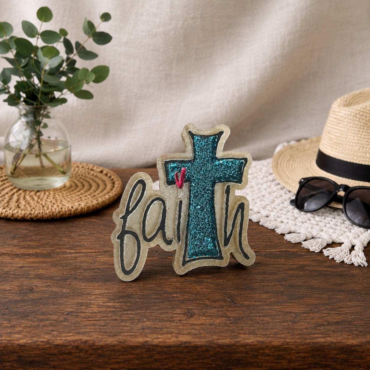 A decorative car freshie spelling 'faith' with a glittery blue cross and a small pink heart, styled on a wooden table with a straw hat, sunglasses, a glass vase with greenery, and a cream fabric backdrop.
