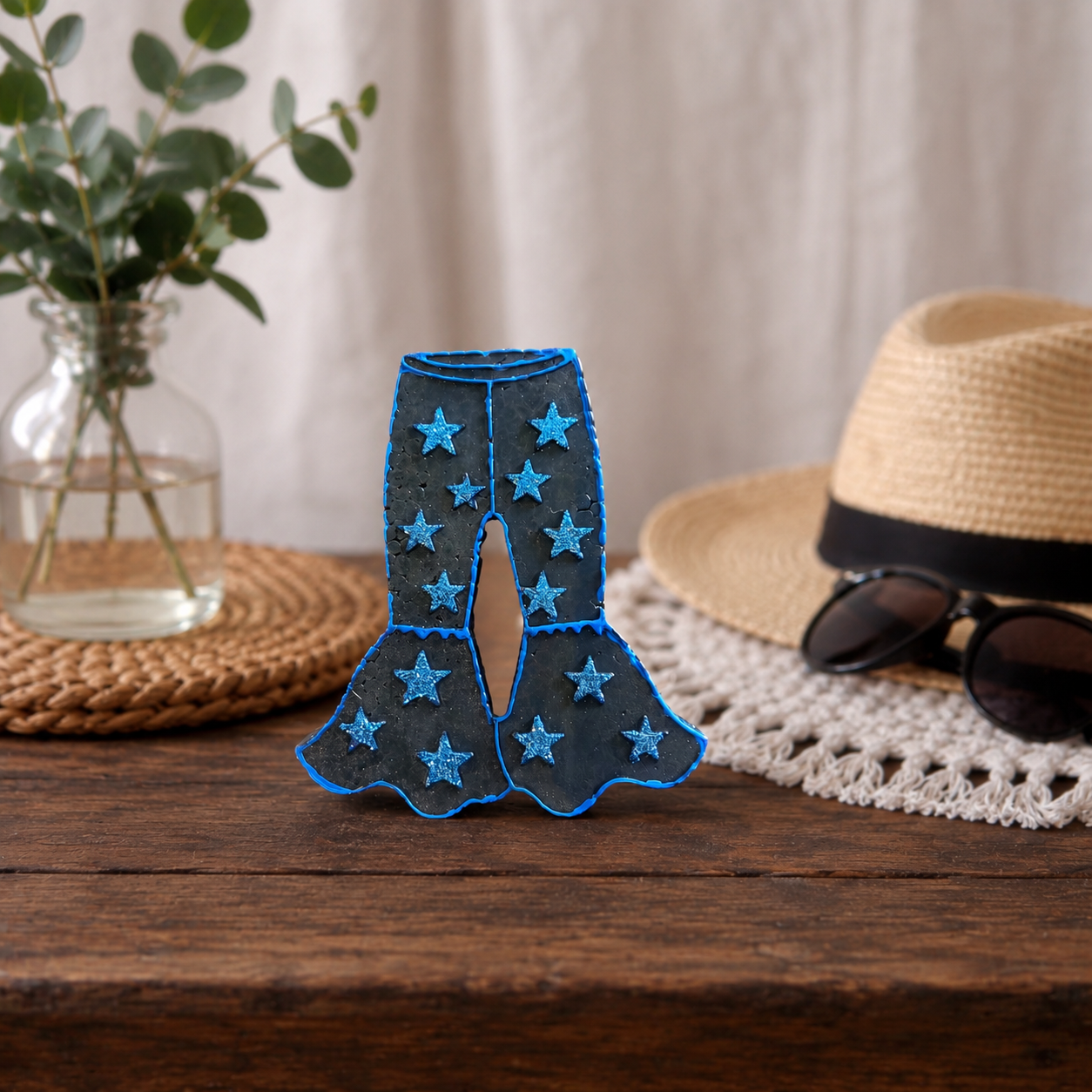 A pair of black denim bell-bottom pants with bright blue glitter star patterns and blue stitched trim, displayed on a rustic wooden table with a straw hat, sunglasses, and a glass vase in the background.