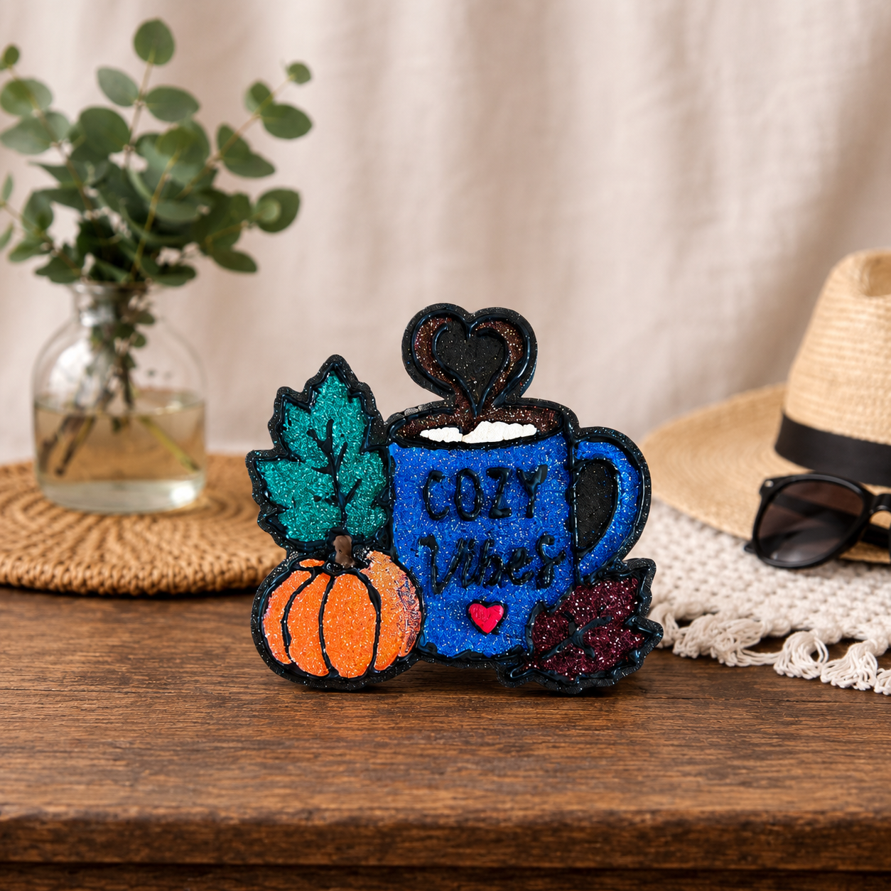 A decorative car freshie shaped like a blue mug with the words “COZY vibes,” surrounded by a pumpkin, teal leaf, dark berry leaf, and a heart-shaped steam detail, displayed on a wooden surface with a hat, sunglasses, and plant in the background.
