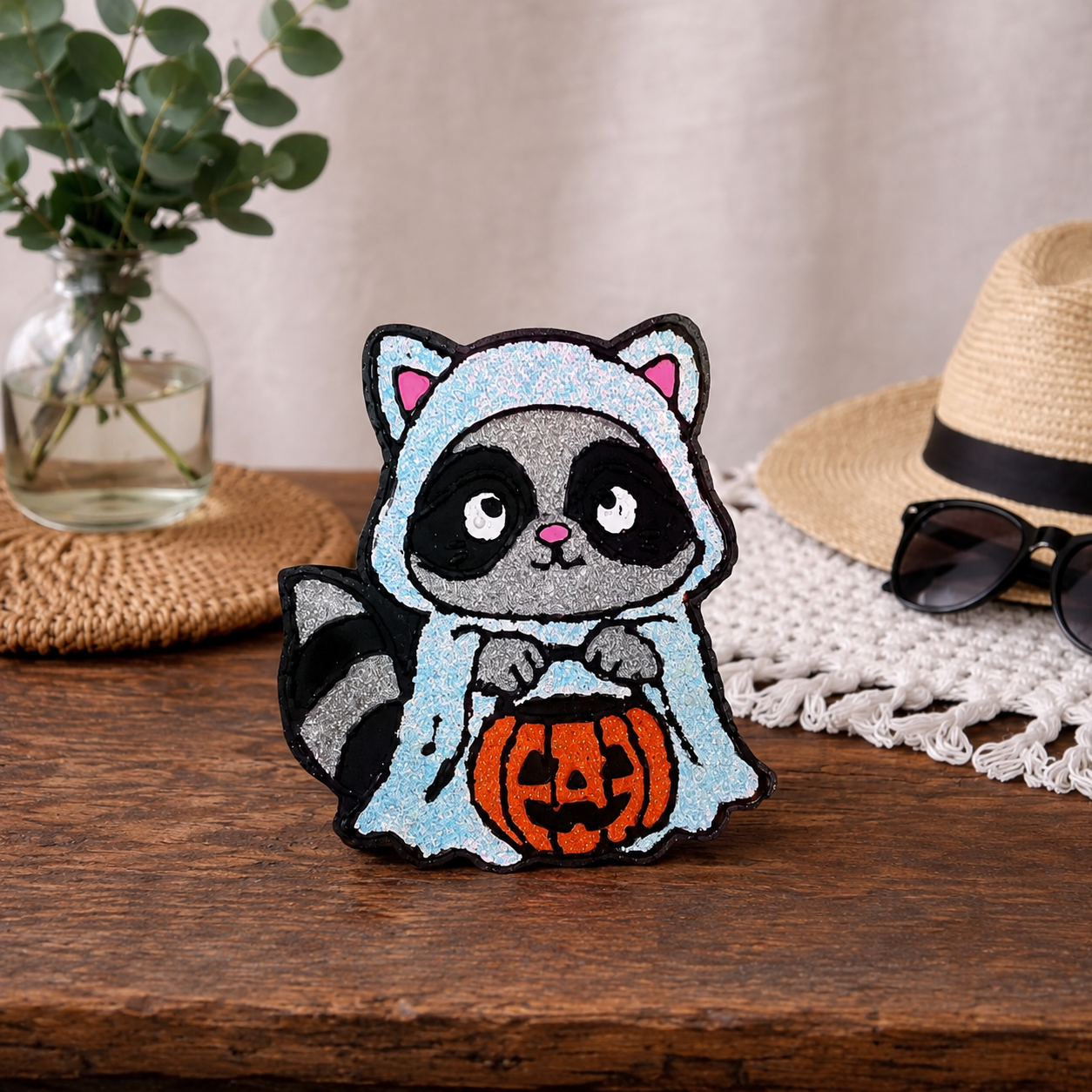 A cute raccoon car freshie dressed in a white glittered ghost costume and holding an orange glittered jack-o'-lantern, displayed on a wooden table with a straw hat, sunglasses, and a glass vase in the background.
