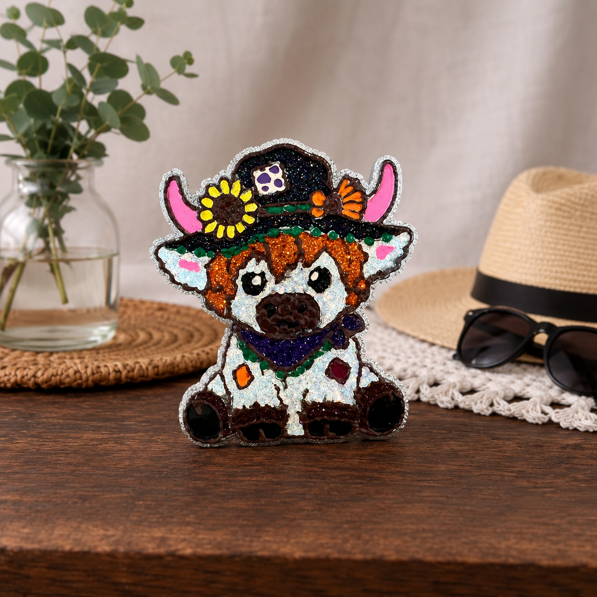 A cow-themed car freshie with a black glitter hat, pink horns, orange hair, and a purple scarf, displayed on a wooden table with a straw hat, sunglasses, and a plant in the background.
