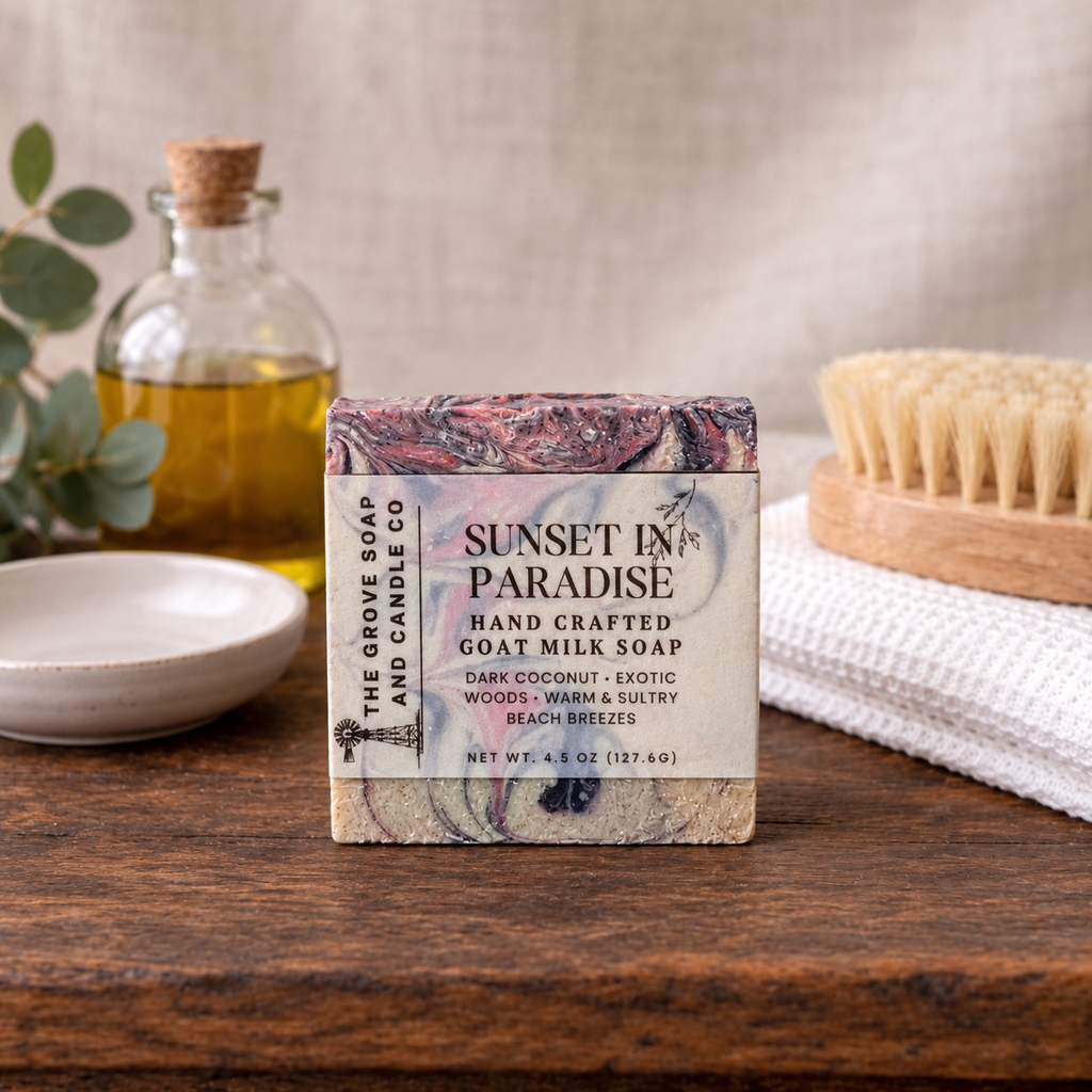 A bar of handmade goat milk soap from The Grove Soap and Candle Co. labeled 'Sunset in Paradise,' displayed on a rustic wooden table with a glass bottle of oil, a small dish, and a wooden brush in the background.