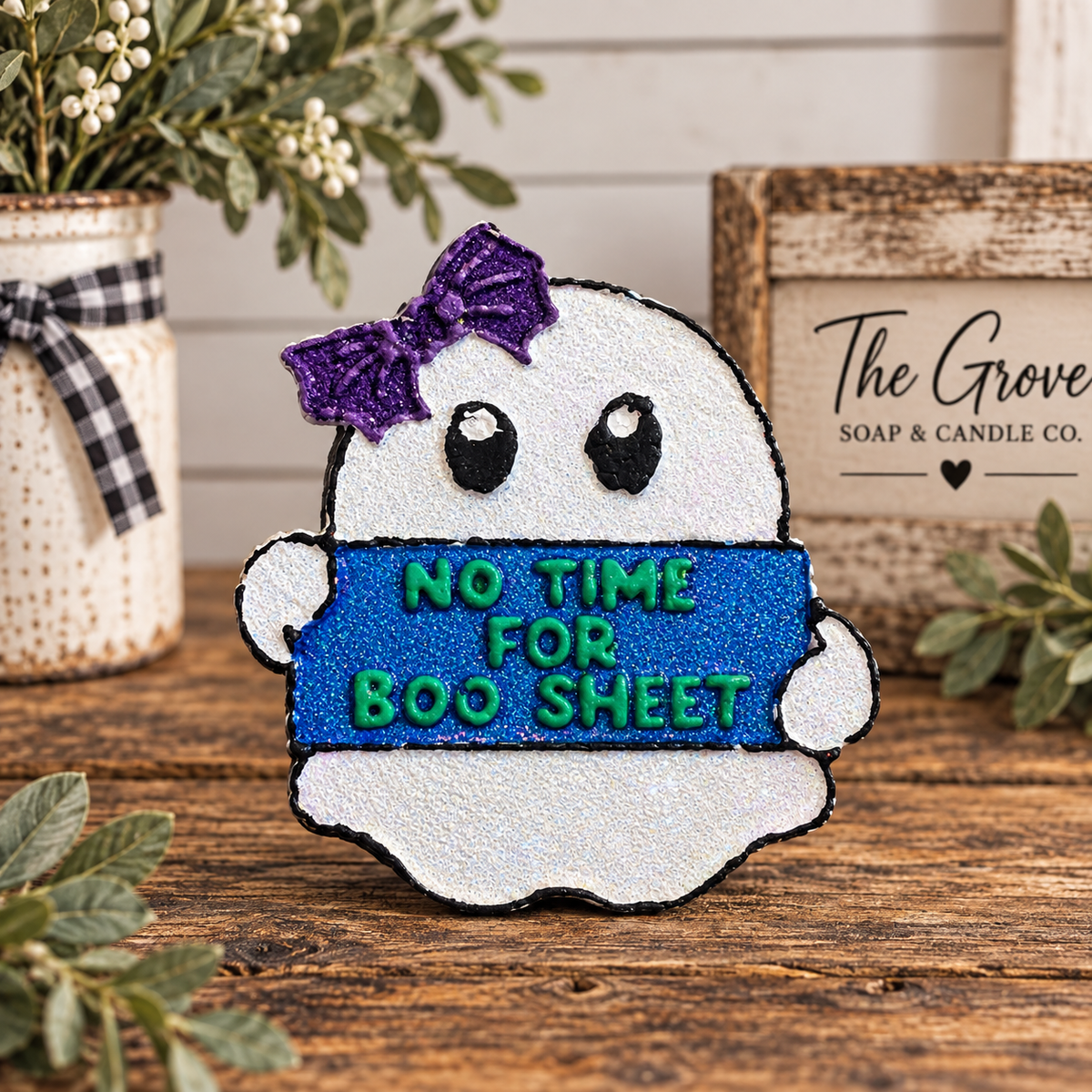 A ghost-shaped Halloween car freshie with a purple bow and the phrase 'NO TIME FOR BOO SHEET' in glittery blue and green, displayed on a wooden surface with decorative plants and a sign reading 'The Grove Soap & Candle Co.'.