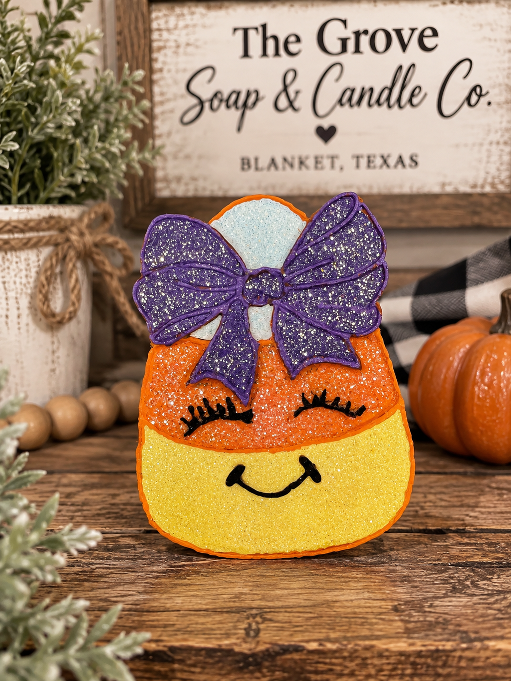 A decorative candy corn freshie with a purple glitter bow, black eyelashes, and a smiling face, displayed on a rustic wooden surface with fall-themed props and a sign reading 'The Grove Soap & Candle Co. Blanket, Texas'.