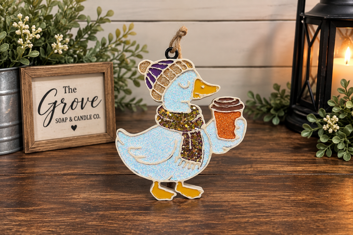 A white glittered duck car freshie with a golden glittered scarf and hat. Holding a coffee cup and sitting on a wooden table. 