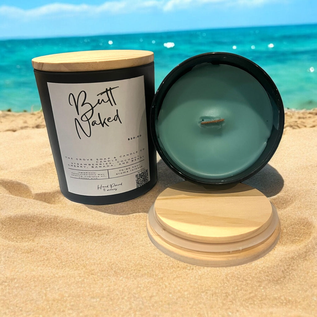 A matte black candle jar with a natural wooden lid, labeled 'Butt Naked', containing a green candle, placed on sand with the ocean in the background.