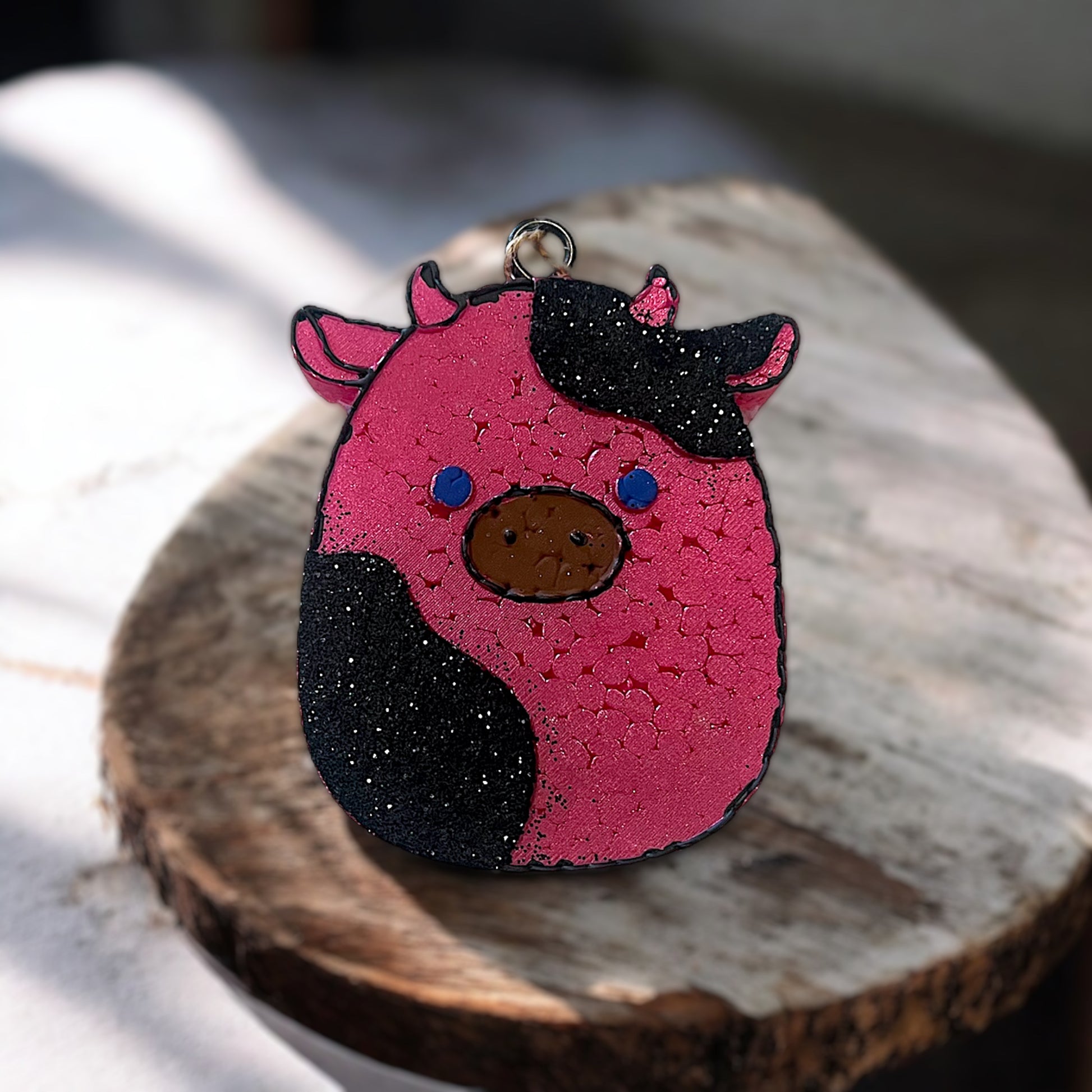 Pink and black glittered car freshie shaped like a cow on a wooden surface