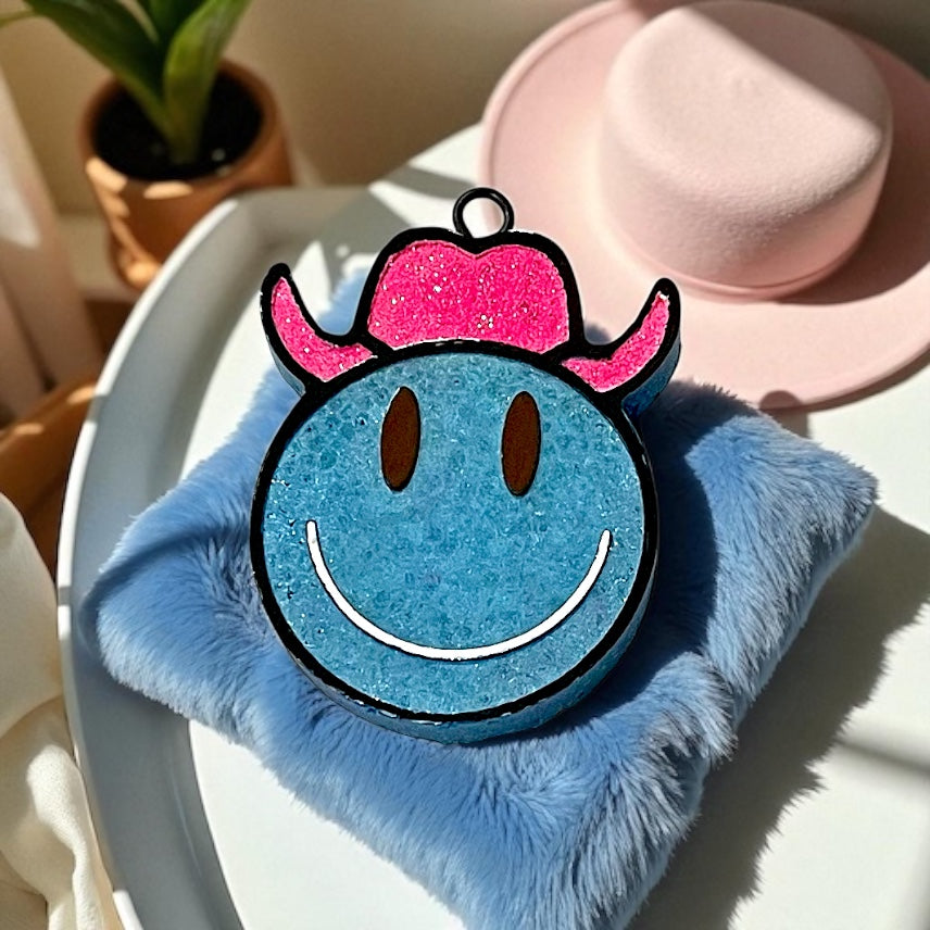 Blue smiley face car freshie with pink glitter cowboy hat on a fluffy blue pillow.