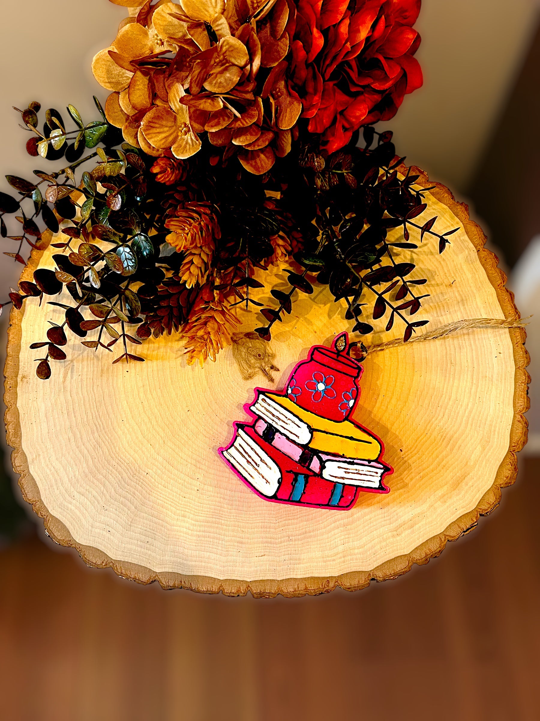 Colorful book-shaped car freshie on a wooden surface with flowers in the background