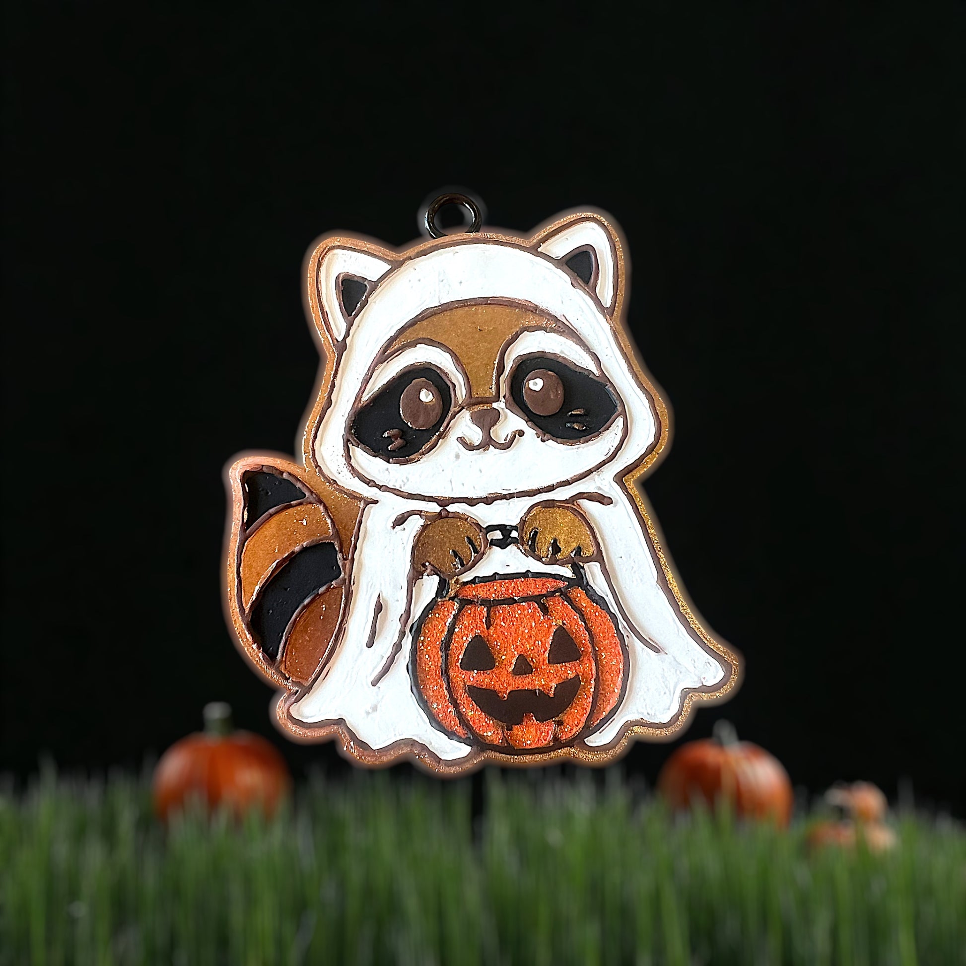 Cute raccoon Halloween car freshie holding a pumpkin on grass with pumpkins in the background.