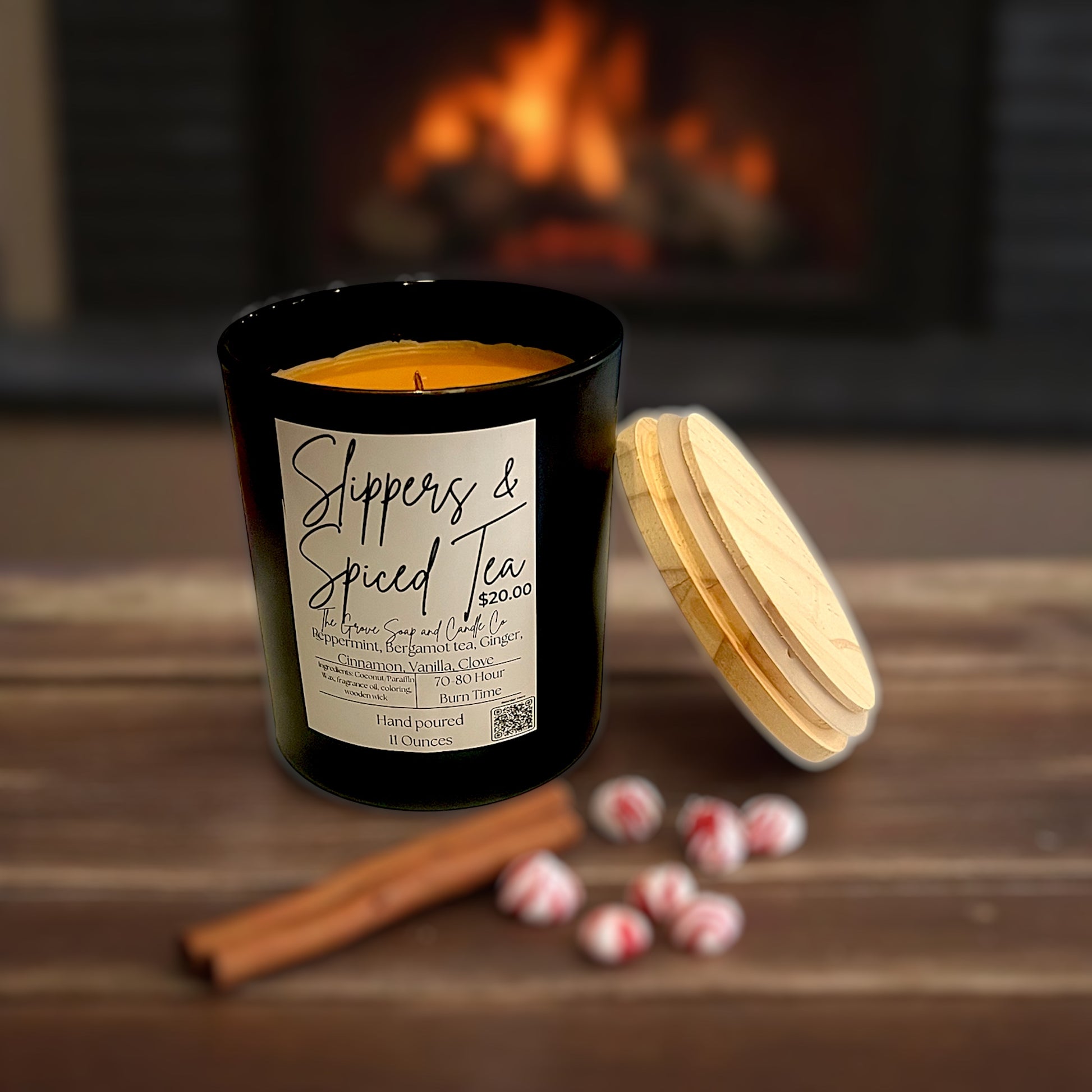 Candle labeled 'Slippers & Spiced Tea' with cinnamon stick and candy on a wooden surface in front of a fireplace.