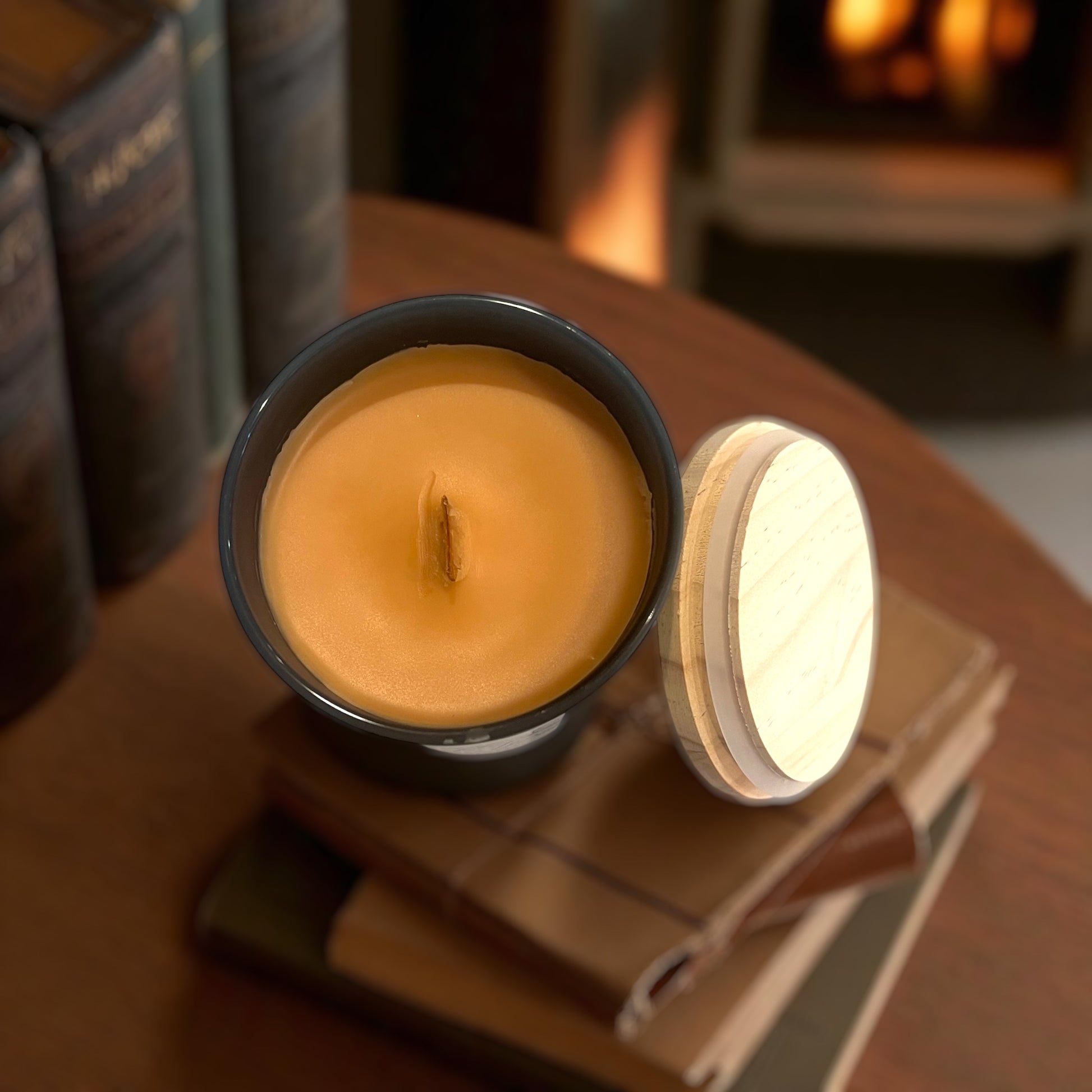 Candle in a black container with a wooden lid on a stack of books, fireplace in the background. Shows the woodeen wick and smooth blend of paraffin and coconut wax. 