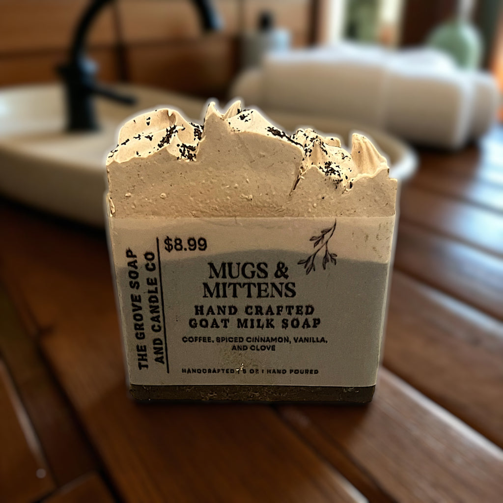 Bar of Mugs & MIttens goat milk soap with a label on a wooden surface, in a bathroom setting.