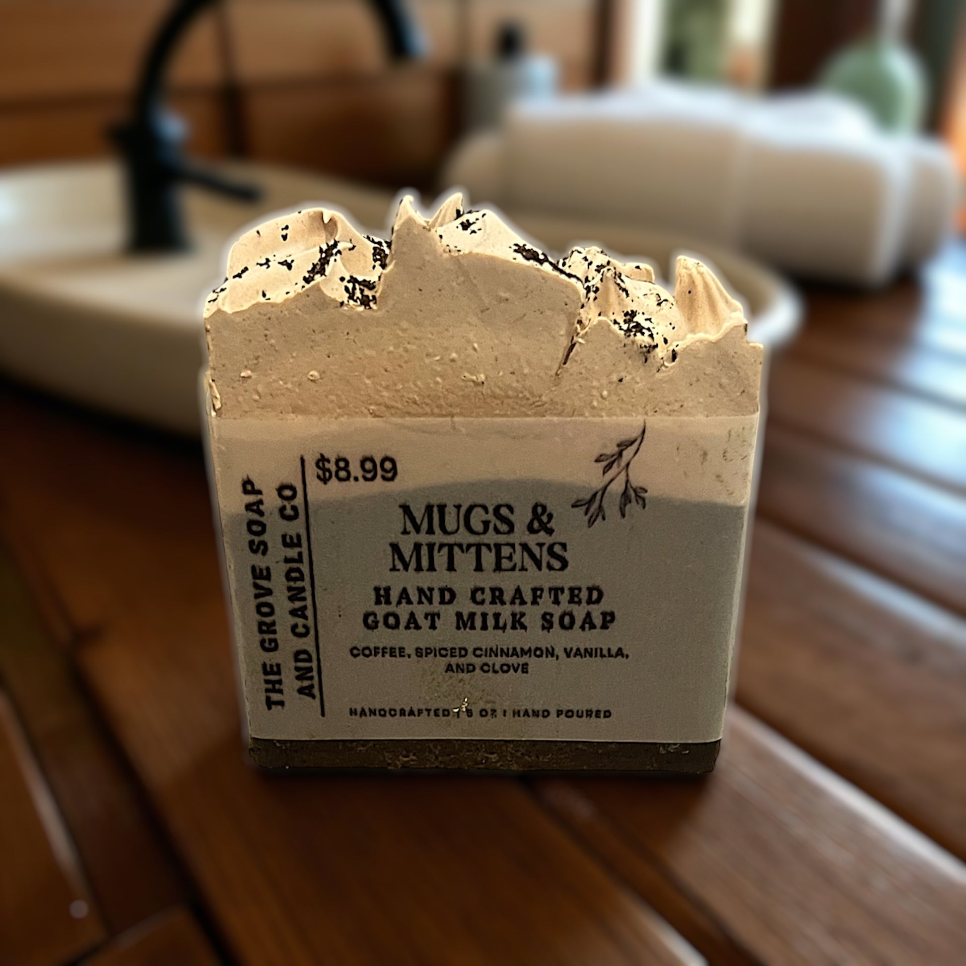 Bar of Mugs & MIttens goat milk soap with a label on a wooden surface, in a bathroom setting.