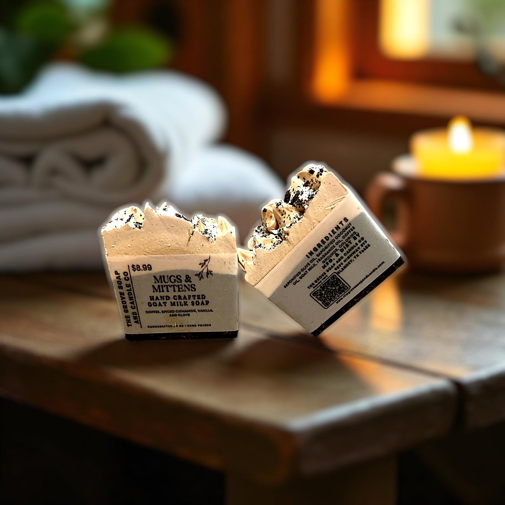 Two small bars of 'Mugs & Mittens' bar soap on a wooden surface with a blurred background. Depicts the labeling for The Grove Soap and Candle with ingredients. 