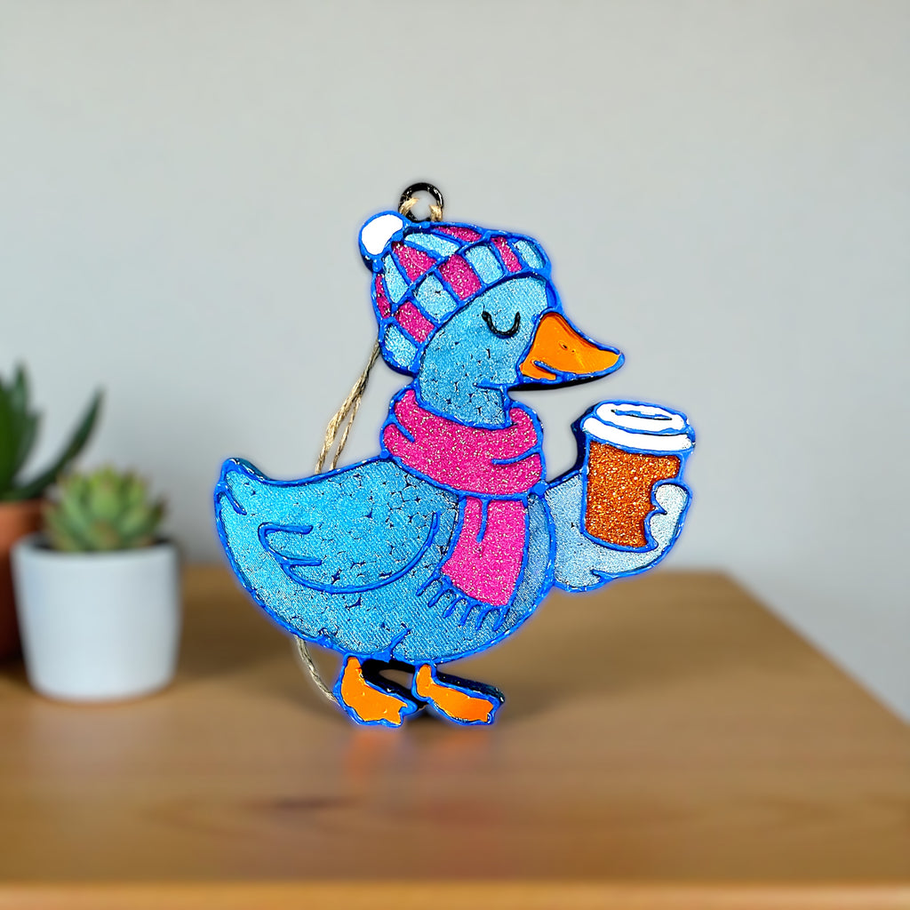Colorful duck car freshie in blue and pink holding a coffee cup on a wooden surface with plants in the background