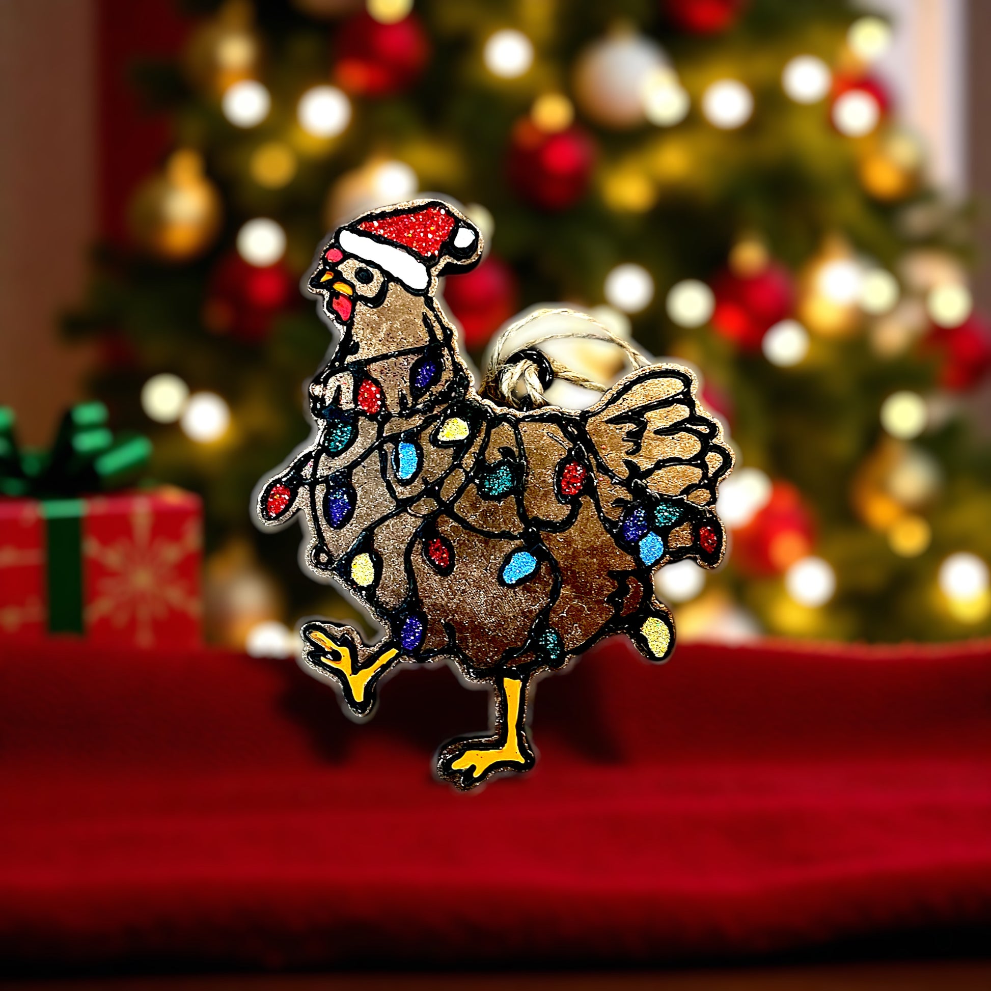 Decorative Christmas chicken car freshie with lights on a red surface with a blurred Christmas tree in the background.