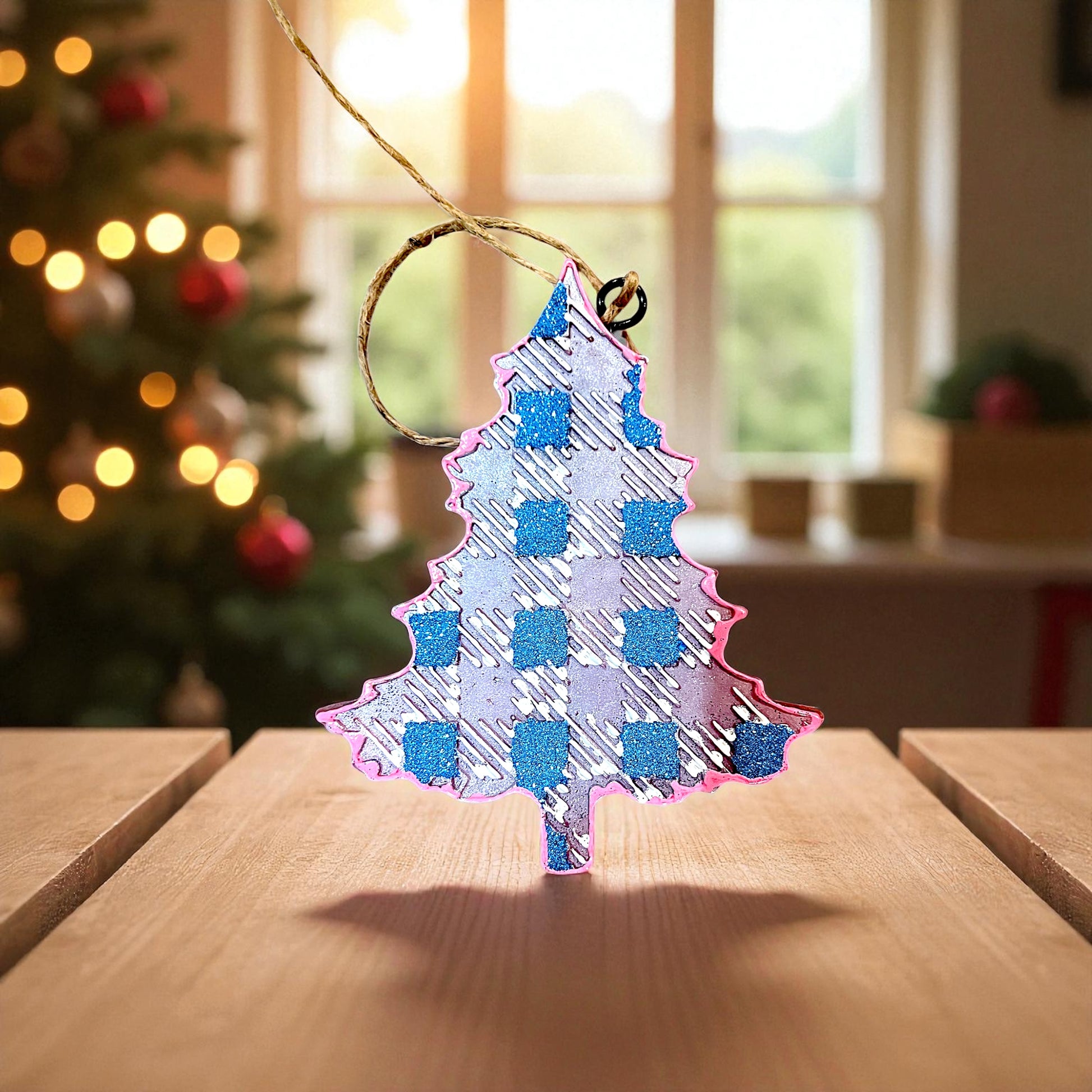 Decorative Christmas tree car freshie with blue and pink plaid pattern on a wooden surface, blurred Christmas tree in the background.