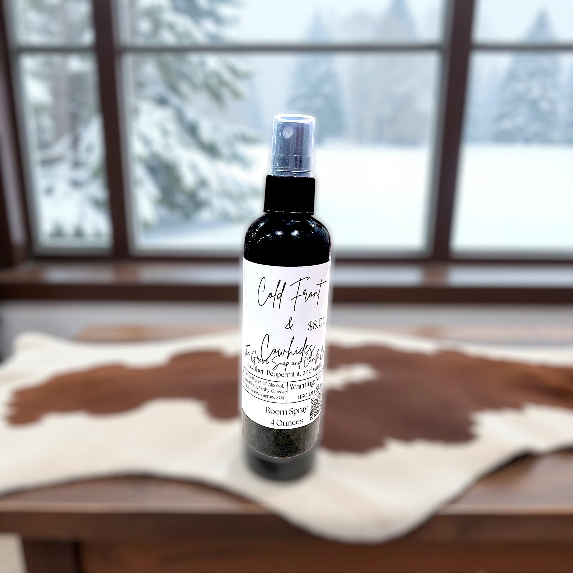 Bottle of room spray with the label Cold Front & Cowhides on a wooden surface with a snowy window background