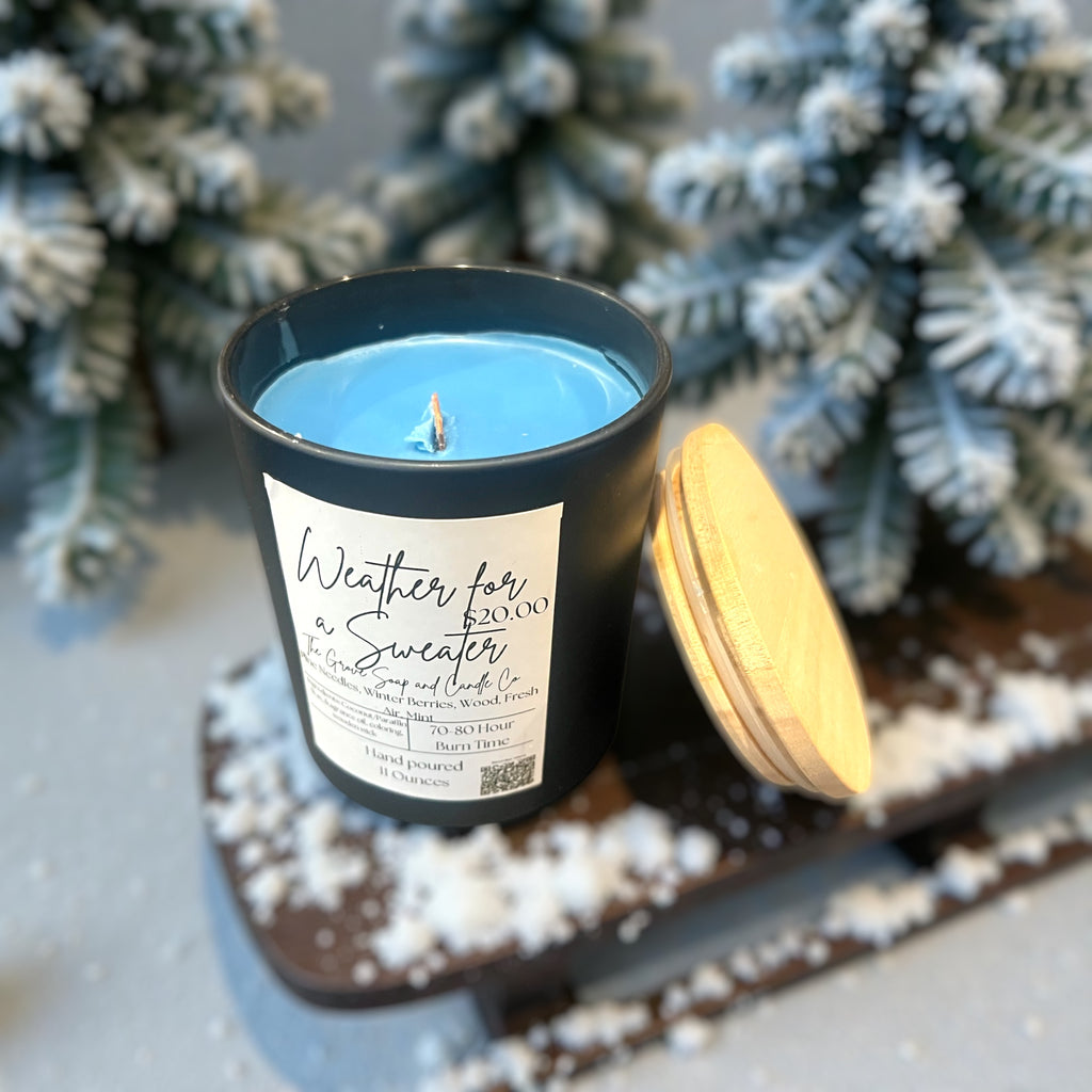 Blue candle Weather for a Sweater in a black container with a wooden wick with a label, surrounded by decorative elements