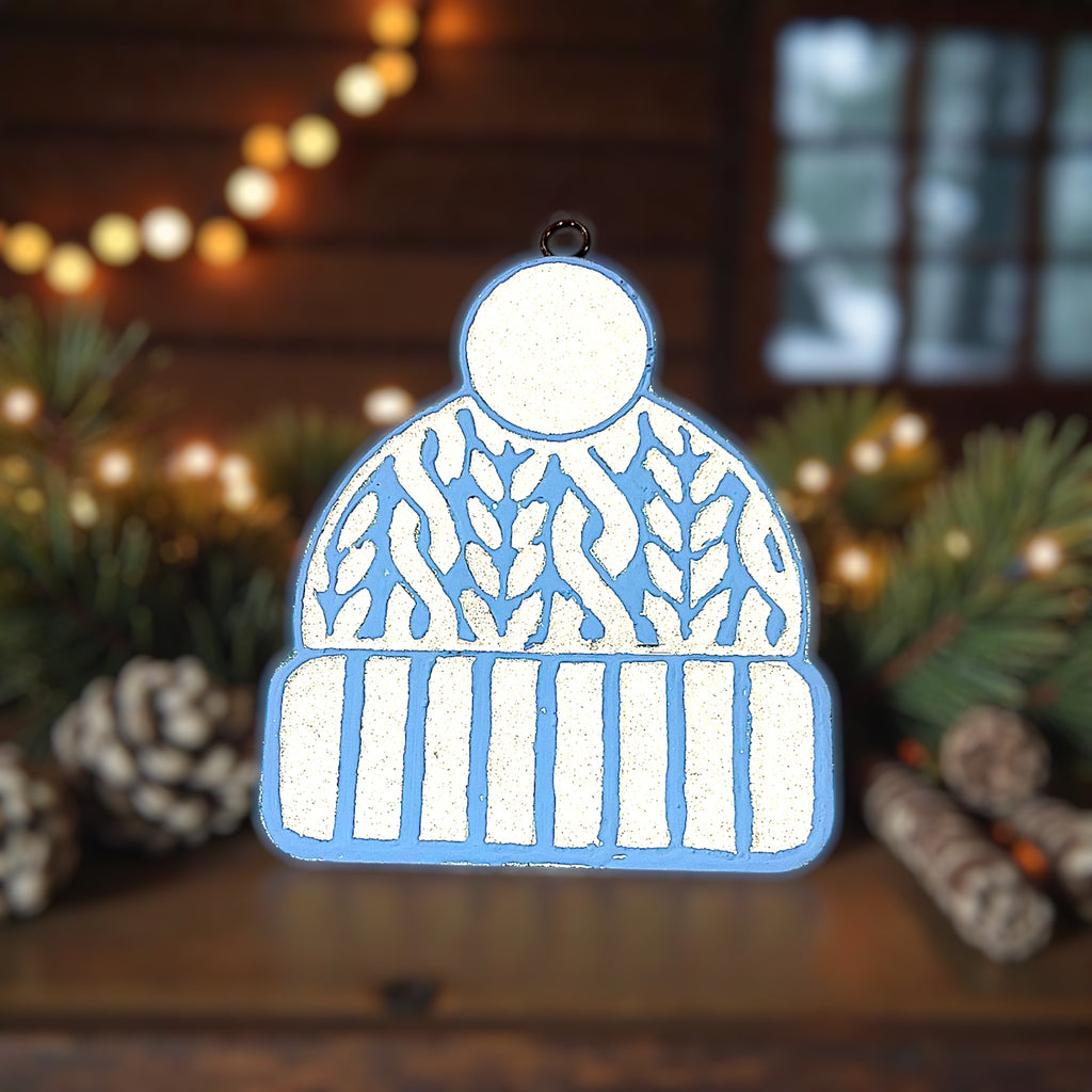 Decorative car freshie shaped like a beanie with a glittered pattern, set against a festive background with lights and greenery.