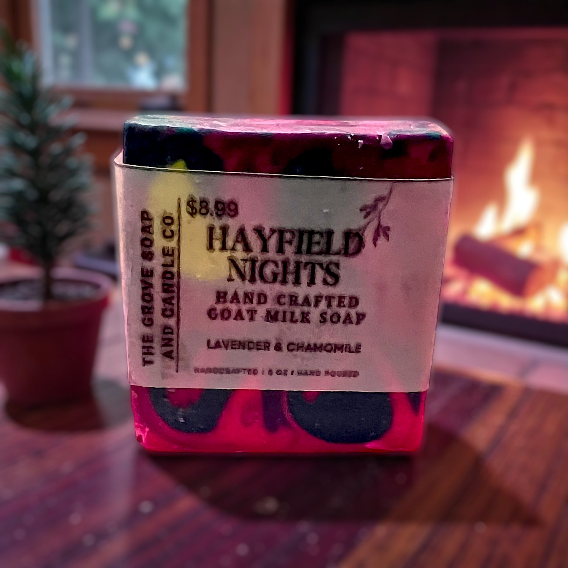 Handcrafted goat milk soap bar with packaging in front of a fireplace. shows the name Hayfield Nights