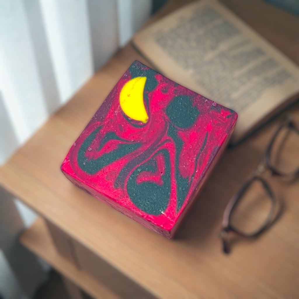 Colorful soap bar with a yellow crescent moon design on a wooden surface next to glasses and a book.